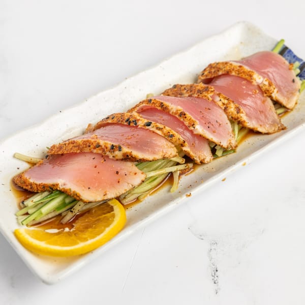 Seared tuna sashimi slices with seasoning, cucumber, and an orange slice on a white rectangular plate.